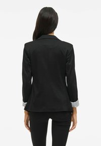 Frau, die mit dem Rücken zur Kamera steht, trägt ein schwarzes Blazer mit gestreiften Ärmeln und schwarze Hose, vor einem schlichten weißen Hintergrund.