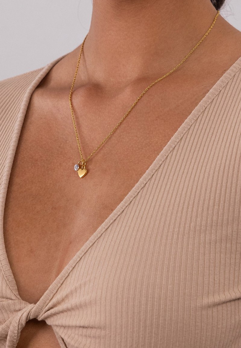 Gouden ketting met hartvormige hanger en kleine transparante edelsteen, gedragen om de hals over een beige ribgebreide top met strik aan de voorkant.