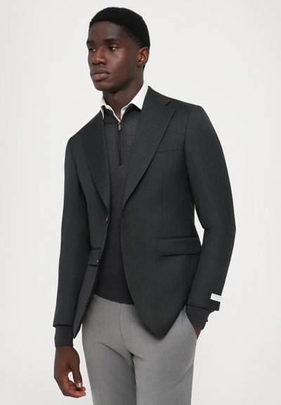 Blazer în dungi gri închis, cu rever în formă de notch, închidere cu un singur nasture, două buzunare frontale, purtat peste un pulover închis la culoare, asortat cu pantaloni gri deschis.