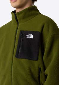 Chaqueta de forro polar de color verde oliva con cuello alto, que cuenta con un bolsillo en el pecho con cremallera negra y detalles negros en los bolsillos laterales inferiores.