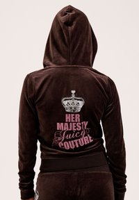 Person trägt einen braunen Velours-Hoodie mit hochgeklapptem Kapuzen, auf dessen Rückseite eine silberne Krone und der pinke Text "Ihre Majestät Juicy Couture" auf einem schlichten Hintergrund zu sehen sind.