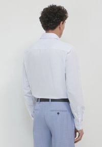 Homme avec des cheveux bouclés portant une chemise habillée rayée blanche rentrée dans un pantalon habillé bleu clair avec une ceinture noire, faisant face à un mur blanc.