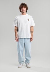 White cotton t-shirt with brown graphic, relaxed fit. Paired with light blue denim pants and white sneakers. Simple, casual outfit.