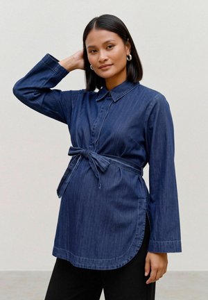 Une femme pose, une main derrière la tête, vêtue d’une tunique en jean bleu foncé ceinturée, sur un fond uni et clair.