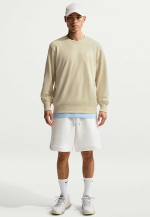 Ung man klädd i beige sweatshirt, vita shorts, vit Nike-keps, vita strumpor och vita sneakers står mot en enfärgad bakgrund.