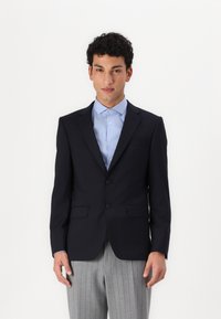 Ναυτικό σακάκι με κανονικά πέτα, δύο κουμπιά και πλαϊνές τσέπες. Συνδυασμένο με ένα ανοιχτό μπλε πουκάμισο και γκρι ριγωτό παντελόνι.