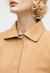 Γυναίκα με κοντά σγουρά ξανθά μαλλιά φοράει μεγάλο αφηρημένο χρυσό σκουλαρίκι και μπεζ σουέτ παλτό με γιακά.