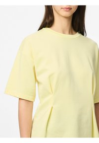 T-shirt jaune clair à manches courtes, col rond, fabriqué en coton doux, présentant des plis subtils sur les côtés pour une forme ajoutée.