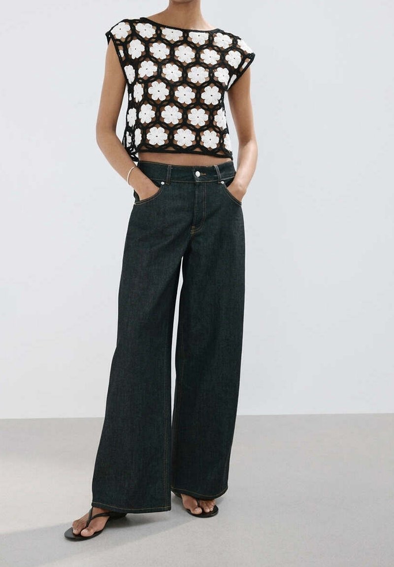 Femme portant un jean noir à jambes larges et un crop top noir en crochet avec des motifs floraux blancs ainsi que des sandales noires.