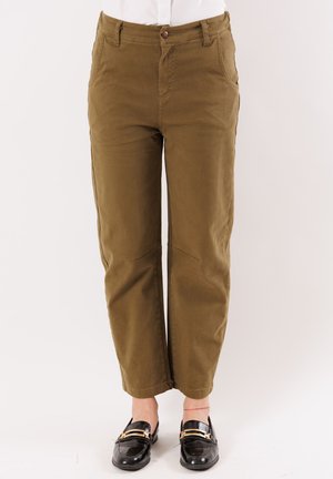 Pantaloni marroni a gamba dritta indossati con mocassini neri e una camicia bianca, mostrati dalla vita ai piedi su uno sfondo chiaro e uniforme.