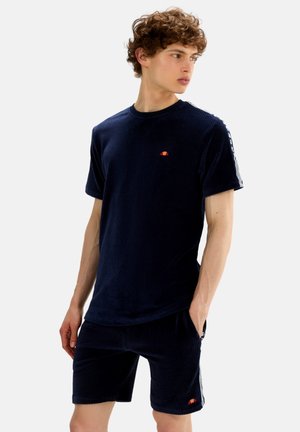 Jonge man in donkerblauw corduroy T-shirt en bijpassende korte broek met witte streepdetails, kijkt naar links met één hand in zijn zak.