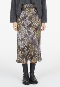Frau in einem Midi-Rock mit Schlangenprint, schwarzen klobigen Stiefeln und einem dunkelgrauen flauschigen Cardigan, die vor einem schlichten Hintergrund steht.