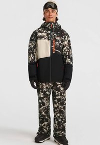 Skijas in zwart, wit en camouflagepatronen met oranje accenten; gecombineerd met camouflagetrouwen broek; handschoenen en skibril inbegrepen.