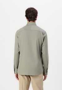 Chemise à manches longues, couleur vert olive, avec un col évasé, un empiècement au dos, une texture lisse et des poignets boutonnés ; associée à un pantalon beige.