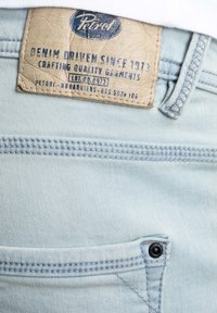 Světle modrá deninová tkanina s koženou nášivkou, na které je vyraženo "Petrol" a "Denim Driven Since 1973." Obsahuje kontrastní švy a nýtek na kapsách.