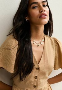 Robe brune texturée avec des manches bouffantes courtes et un décolleté en V profond, présentant trois boutons sur le devant et un collier en chaîne superposé.