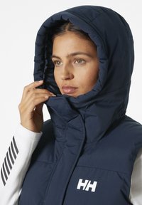 Giacca puffer con cappuccio di colore blu navy, caratterizzata da un design trapuntato, una texture liscia e un logo prominente sul petto. Inquadratura ravvicinata su viso e mani.