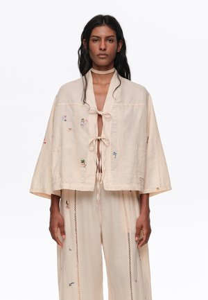 Mujer con top beige suelto con lazos delanteros y diseños bordados sutiles, combinado con pantalones de pierna ancha a juego.