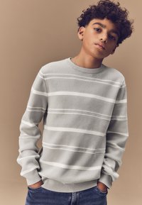 Maglione grigio a righe con bande orizzontali bianche, collo rotondo, polsini a coste, vestibilità rilassata, indossato sopra jeans scuri. Materiale in maglia morbida.