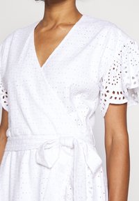 Abito avvolgente bianco in cotone testurizzato con dettagli ad occhio, maniche a sbuffo corte e una cintura legata in vita.