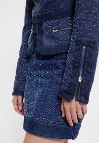 Komplet jeans jakna in krilo v temno modri barvi z madežasto teksturo. Ima sprednji žep z zadrgo in prilegajoče se rokave.