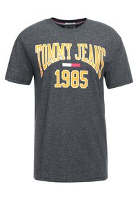 Camiseta de algodón gris con el texto en negrita "TOMMY JEANS" en amarillo y "1985" debajo, con un pequeño acento de logo rojo y blanco. Cuello redondo, mangas cortas.