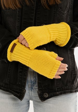 Gelbe gestrickte fingerlose Handschuhe mit gerippter Textur und einem braunen Lederlabel. Kombiniert mit einer schwarzen Jeansjacke.