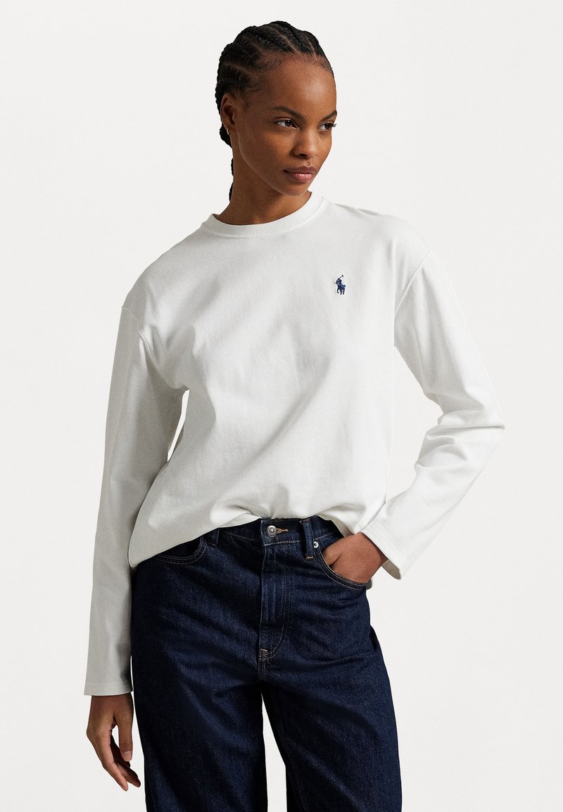 Camisa blanca de manga larga hecha de algodón, con un pequeño emblema azul en el pecho, combinada con jeans de mezclilla oscura.
