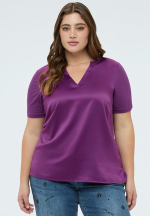 Frau mit langen, welligen Haaren, die ein lila kurzärmeliges V-Ausschnitt-Oberteil und blaue Jeans mit Blumenverzierungen trägt, steht vor einem einfarbigen Hintergrund.