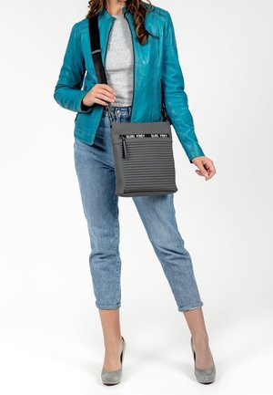 Žena v tyrkysové kožené bundě, šedém topu a spadených modrých džínách drží šedou crossbody tašku s logem "SURI FREY" na popruhu.