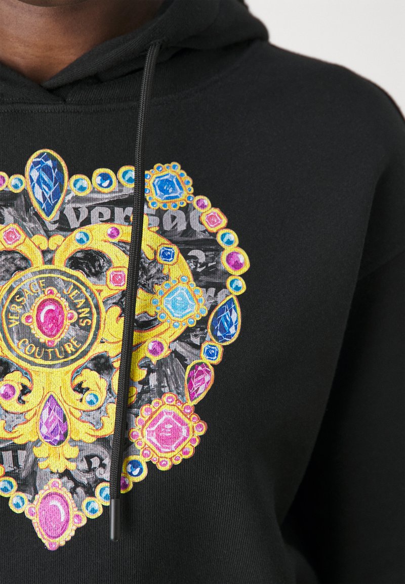 Hoodie noir avec un graphique orné et coloré, comportant des gemmes et des motifs dorés. Le logo indique "VERSACE JEANS COUTURE." Tissu texturé visible.