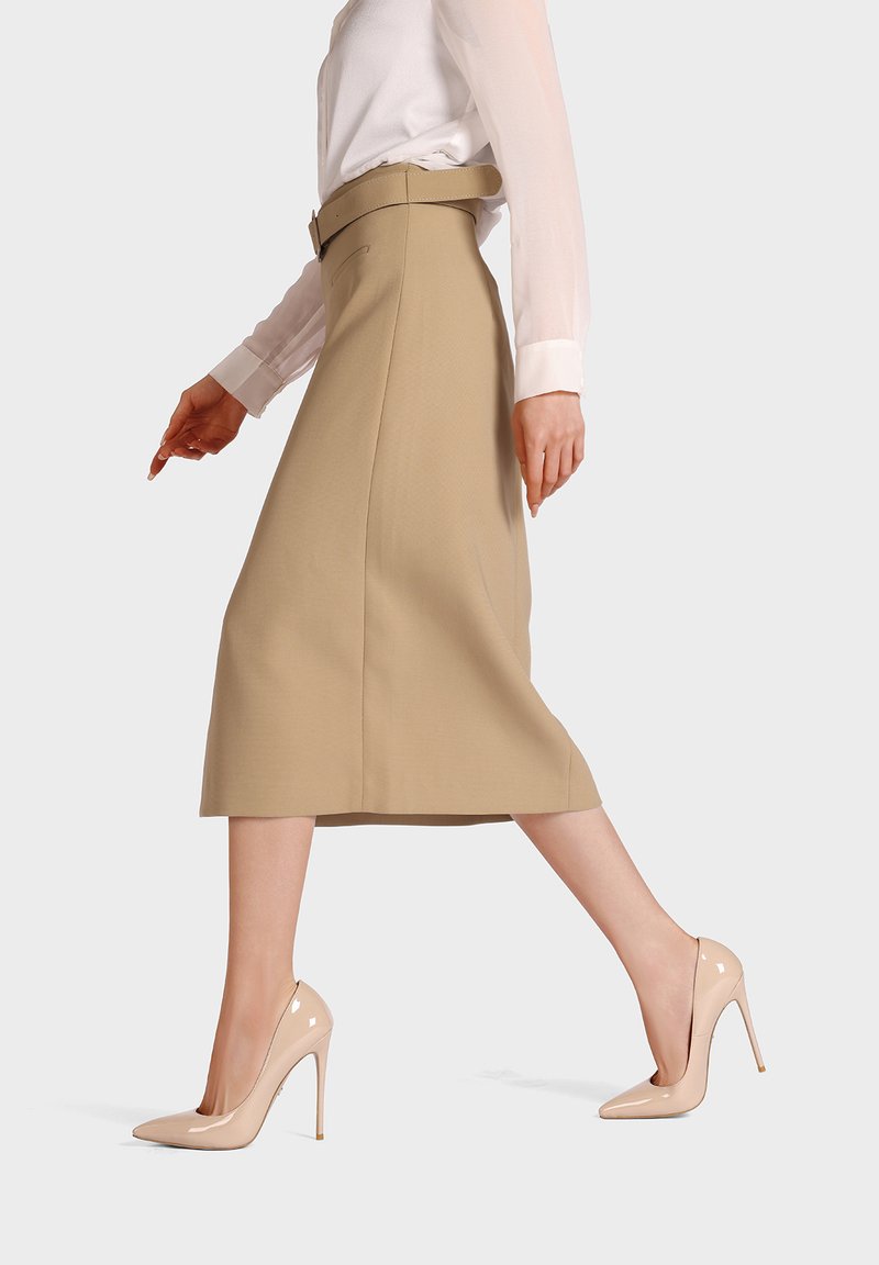 Mujer caminando con falda midi beige con cinturón, blusa blanca de manga larga y zapatos de tacón alto de charol color nude sobre fondo blanco.