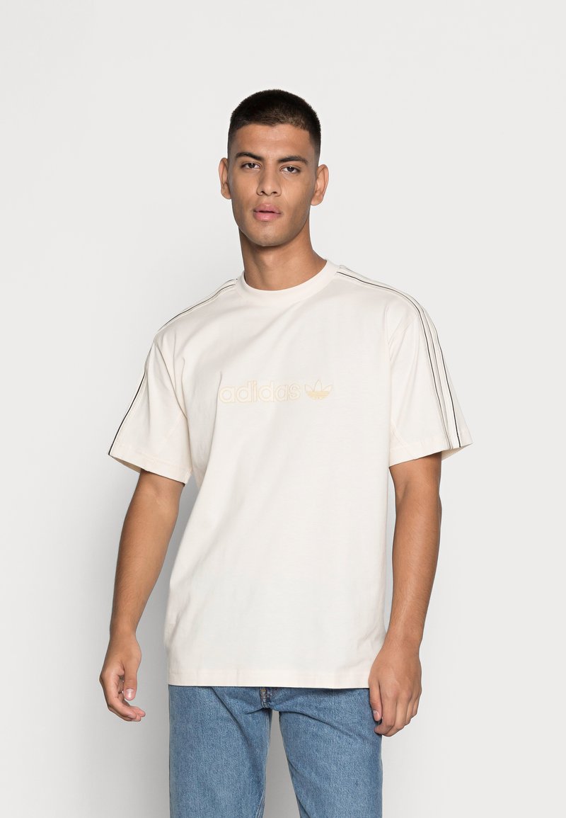 T-shirt cream a maniche corte con logo Adidas dorato, strisce laterali nere e vestibilità rilassata. Indossato con jeans azzurri chiari.