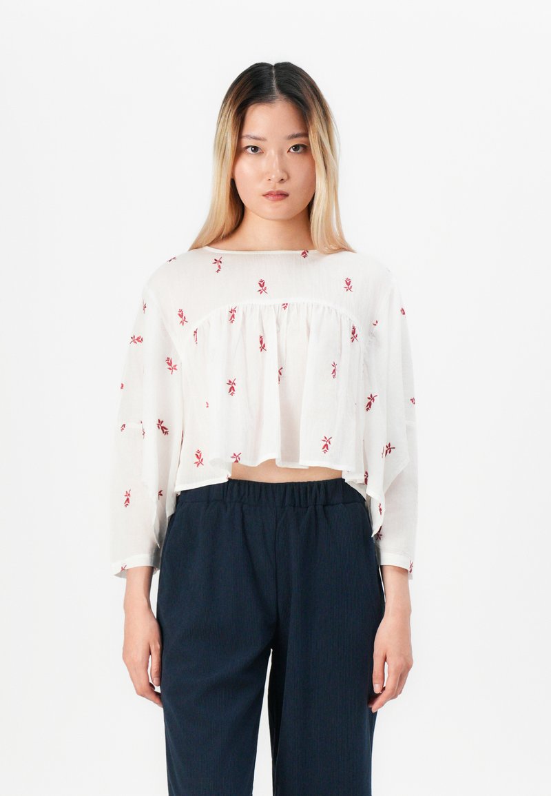 Young woman with long ombre hair wearing a white floral cropped blouse and loose dark navy pants, standing against a plain white background.