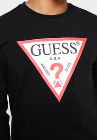 Zwarte lange mouwen shirt met een groot rood en wit driehoekig Guess-logo met een vraagteken en de tekst "U.S.A. Washed Jeans."