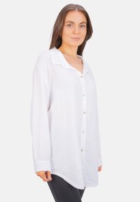 White long-sleeve shirt with a button front, collar, and textured fabric. Curved hem, side slits, and slightly oversized fit.