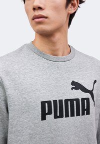Personne portant un sweat-shirt gris clair avec un grand logo Puma noir présentant un puma bondissant sur la poitrine, la tête légèrement tournée vers la gauche.
