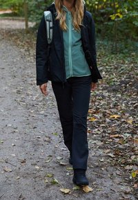 Sort udendørs jakke, grøn fleece zip-up, sorte bukser og solide sorte støvler. Går på en sti med efterårsblade. Rygsæk synlig.