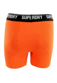 Narančaste bokserice s crnim elastičnim pojasom na kojem se nalazi bijeli logo 'SUPERDRY'. Mekana tekstura, dužina do sredine bedra i prilagođen dizajn.