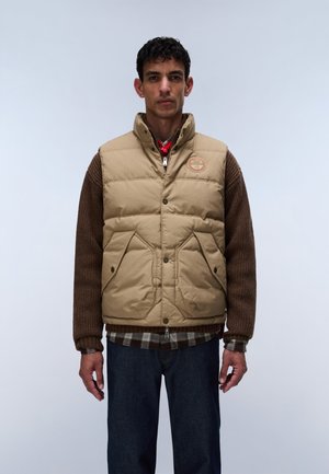 Mand iført beige polstret vest over brun sweater og ternet skjorte, stående mod en ensfarvet lys grå baggrund.