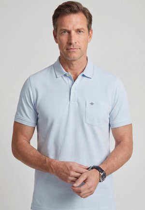 Man draagt een lichtblauw poloshirt met borstzak, stelt zijn polshorloge af, met een neutrale grijze achtergrond.