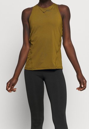 Donna in top atletico senza maniche verde oliva e leggings neri, in piedi con le braccia rilassate ai lati su uno sfondo neutro.