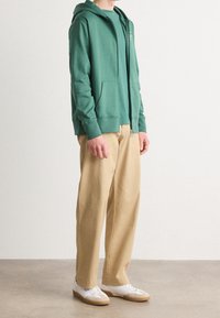 Felpa verde con zip e tasca frontale, abbinata a pantaloni beige e sneakers bianche. Tessuto morbido, vestibilità rilassata e polsini a costine.