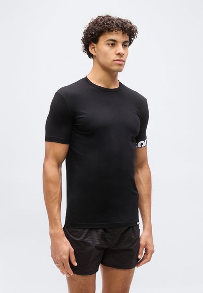 Jeune homme aux cheveux bouclés portant un t-shirt noir ajusté à manches courtes et un short noir à motifs, debout devant un fond clair uni.