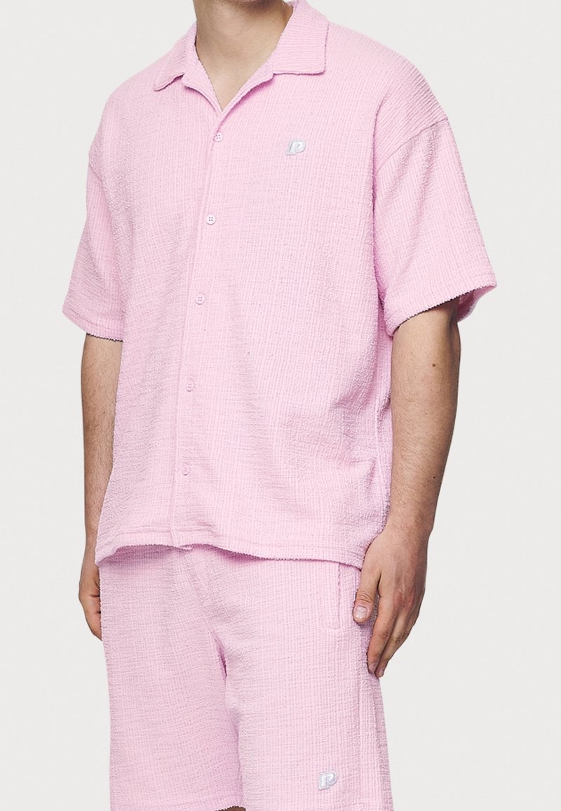 Chemise rose à manches courtes avec boutons et short assorti en tissu texturé. Présente un petit logo brodé sur la poitrine et le short.