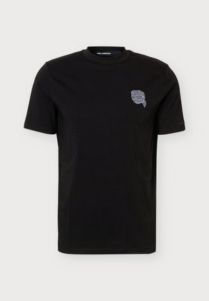 Un homme pose avec un T-shirt noir à manches courtes orné d’un petit emblème sur la poitrine, un jean foncé et des chaussures noires, sur un fond clair uni.
