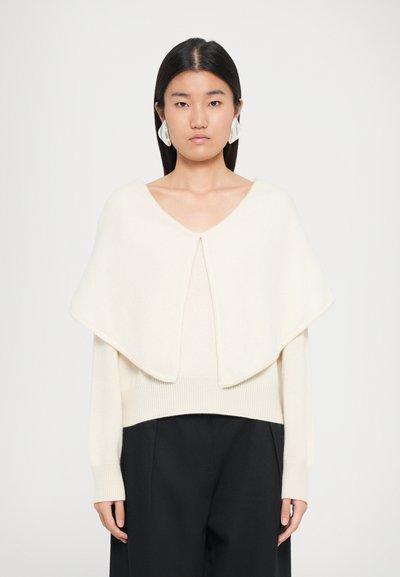 Young woman with straight dark hair wears large white earrings, an off-white knitted top with wide collar, and black trousers against white background.