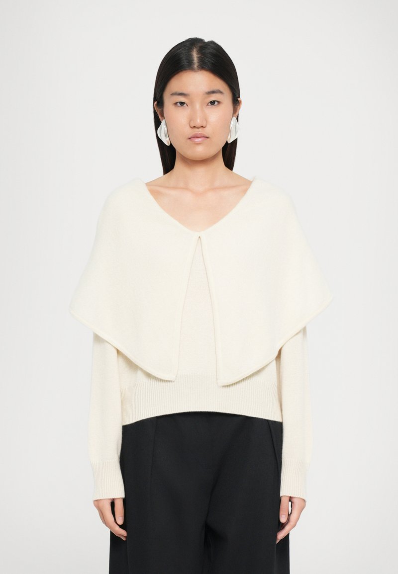Jeune femme aux cheveux raides et foncés portant de grandes boucles d'oreilles blanches, un haut en tricot écru avec un large col, et un pantalon noir sur fond blanc.