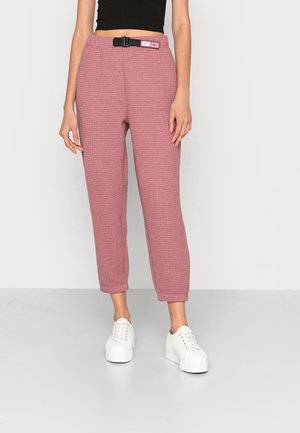 Femme portant un pantalon mauve texturé taille haute avec une ceinture noire à boucle, associée à des baskets à plateforme blanches sur un fond uni.