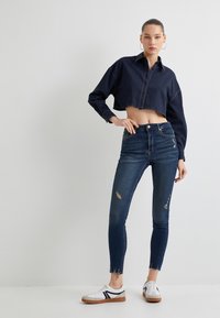Camisa de mezclilla recortada en color marino con cierre de botones, combinada con jeans skinny oscuros desgastados y zapatillas blancas con rayas negras.